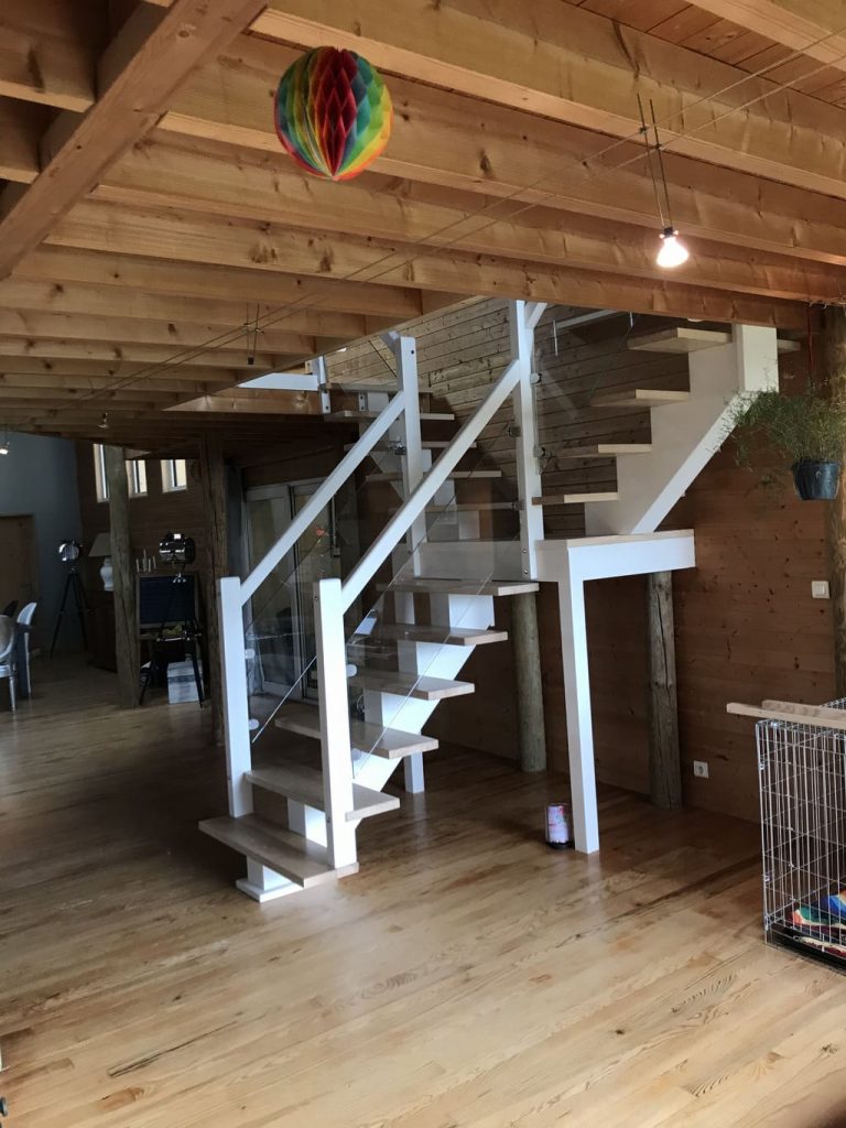 Une pièce avec un escalier central blanc, une rambarde en verre et une cage pour chien.